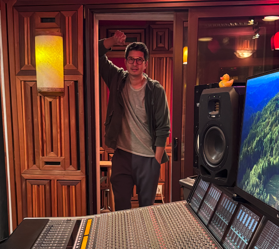 Young man standing in recording studio with wood-paneled walls, raising his right hand, with large speakers and a mixing console and a monitor displaying a forest landscape in the background.