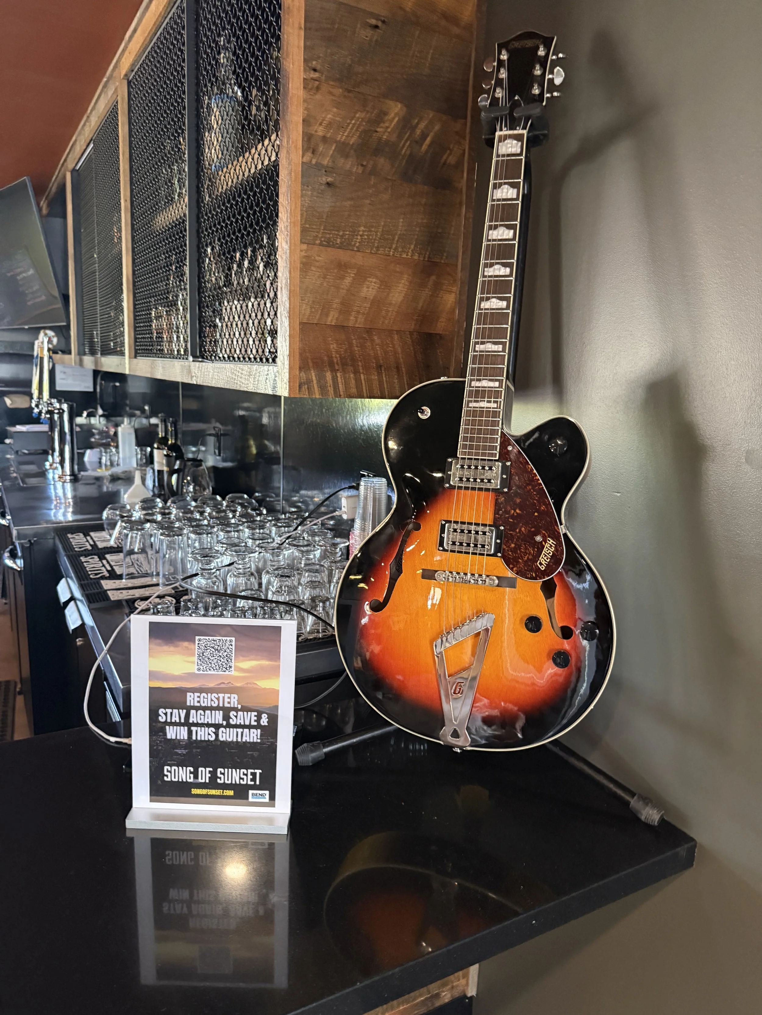 A music stand with a sign promoting a contest to win a guitar, a black and orange semi-hollow electric guitar resting on a stand, a bar counter with glasses, and a wooden shelf in the background.