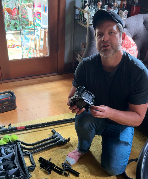 A man kneeling on a yellow rug inside a house, holding a camera with a large lens. There are fishing rods, a gun, and shooting accessories on the rug. Behind him, there is a glass door leading to a patio with toys and outdoor furniture, and a shelf with bottles and decorative items.