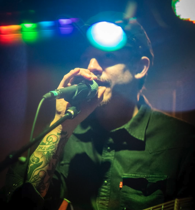 Person singing into a microphone on stage with colorful lights.