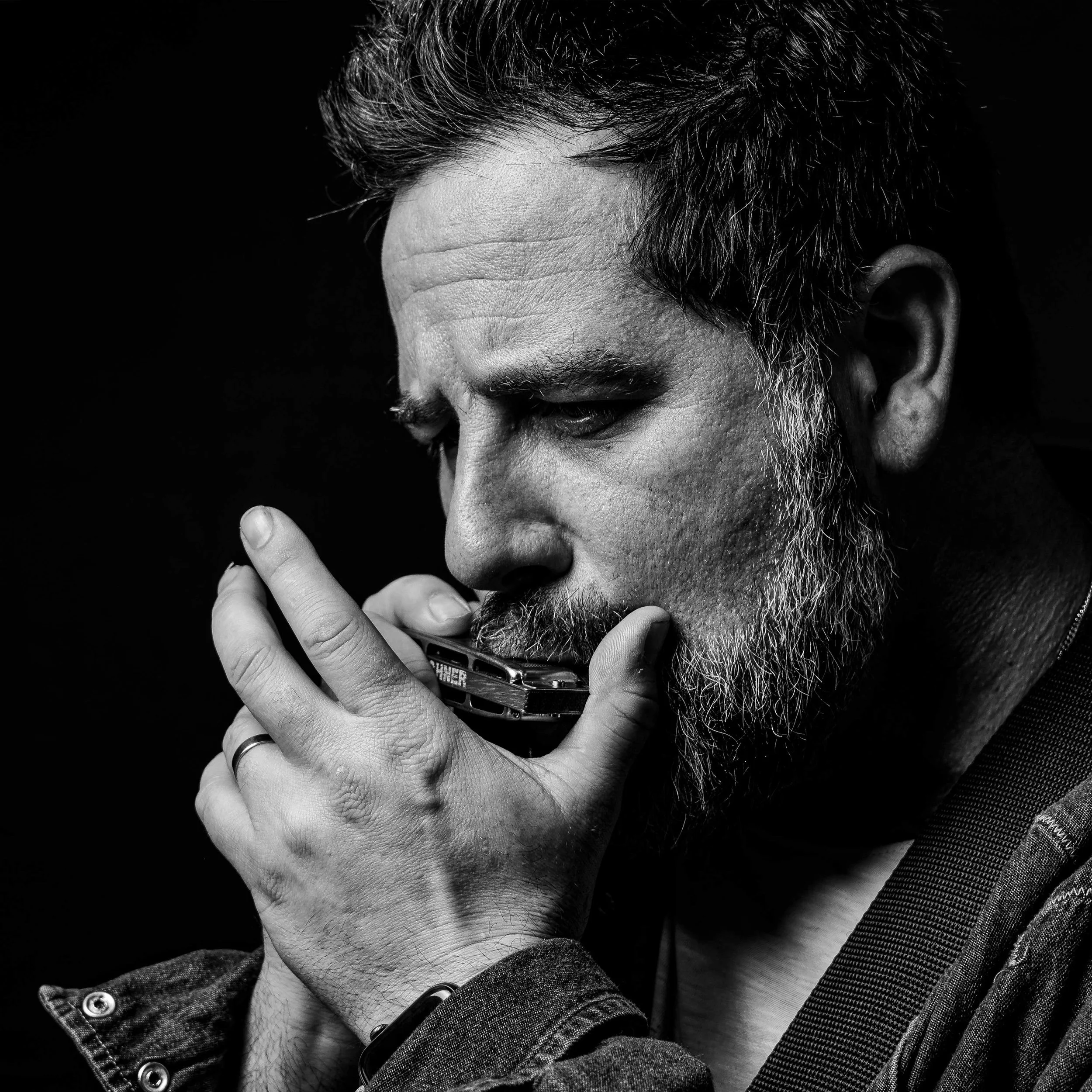 Black and white photograph of a man with a beard and mustache, with short, styled hair, holding a harmonica near his face with his eyes closed.