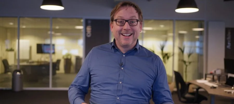 Man met bruin haar en bril in een kantoorruimte, lachend en gekleed in een blauwe blouse.