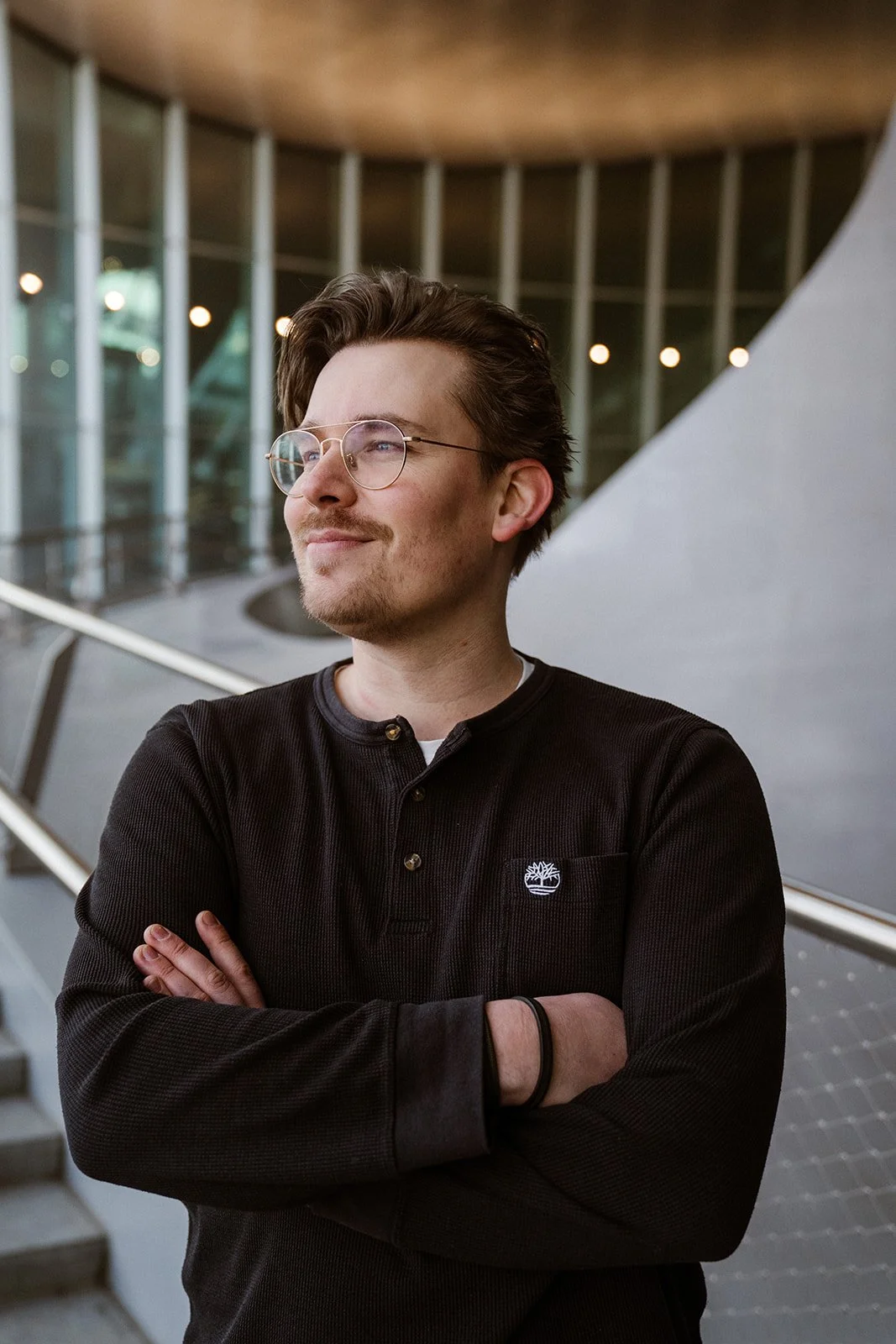 Man met bril, gekleed in een zwarte trui, staand met gekruiste armen voor een moderne, architectonische achtergrond met glas en hout, in een indoor setting.