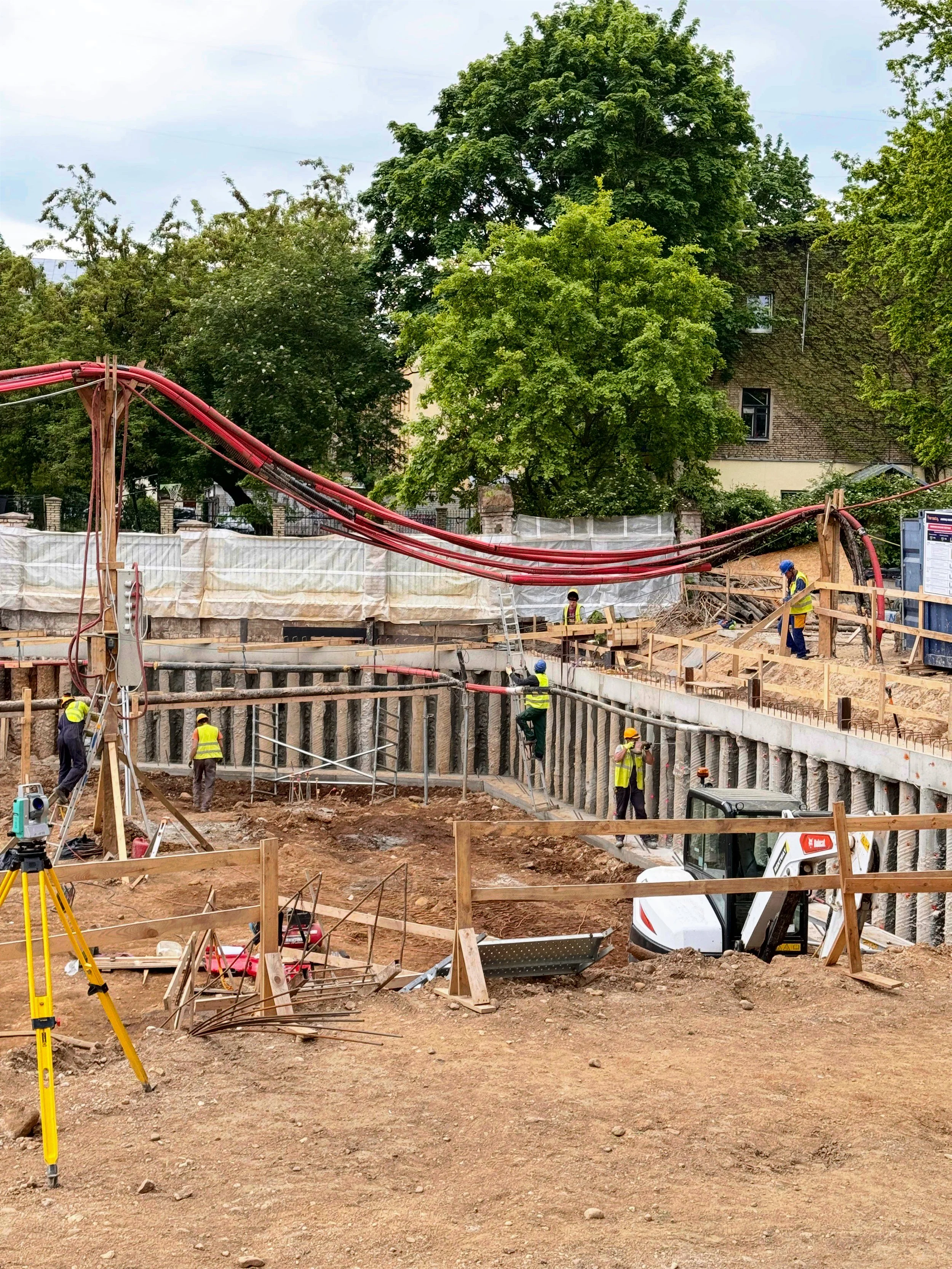 Statybvietė su darbininkais, pastoliais ir pamatų statymu.