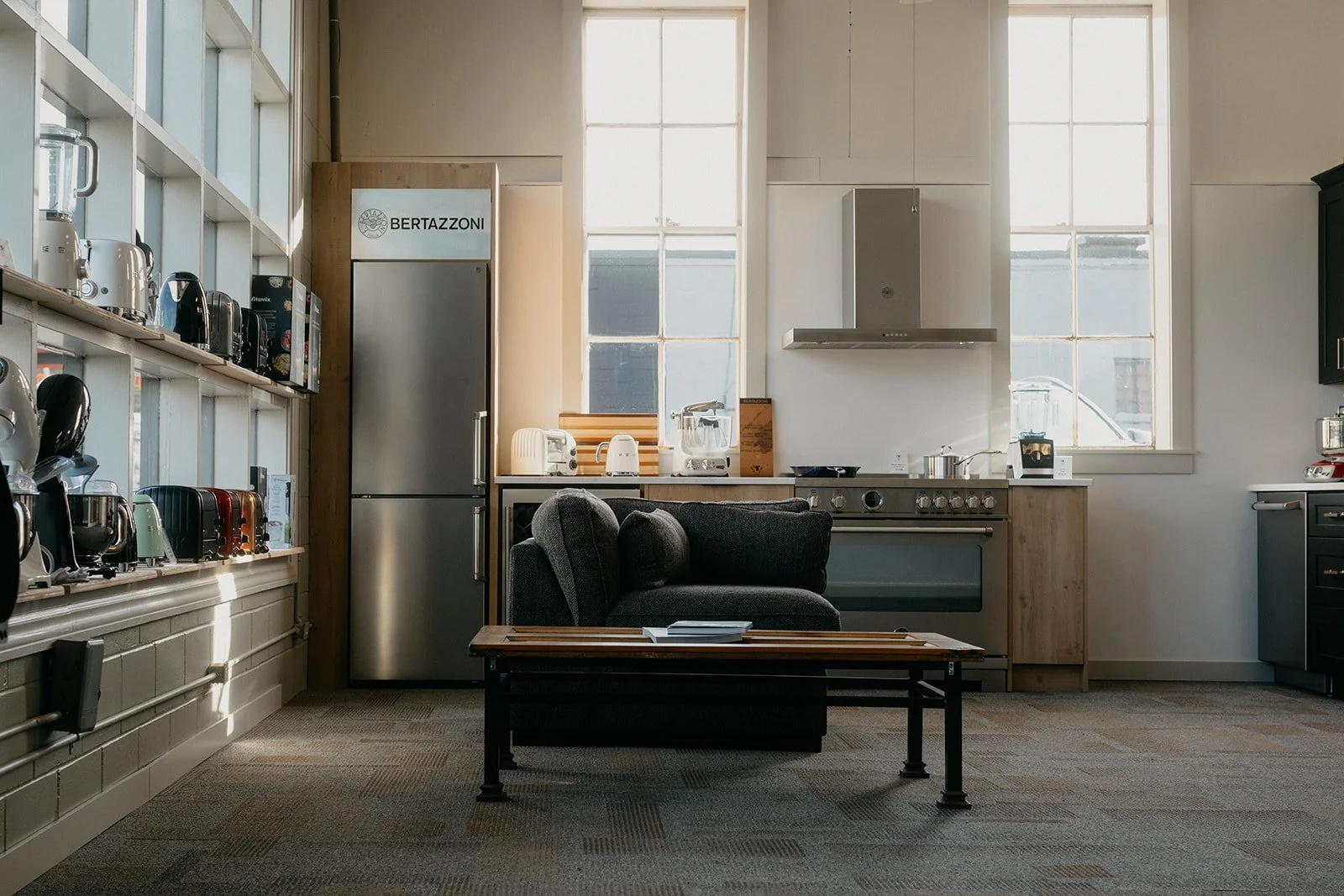 Modern kitchen with a dark sofa and coffee table in the center, stainless steel fridge, a stove, and wooden cabinets, surrounded by large windows letting in natural light.