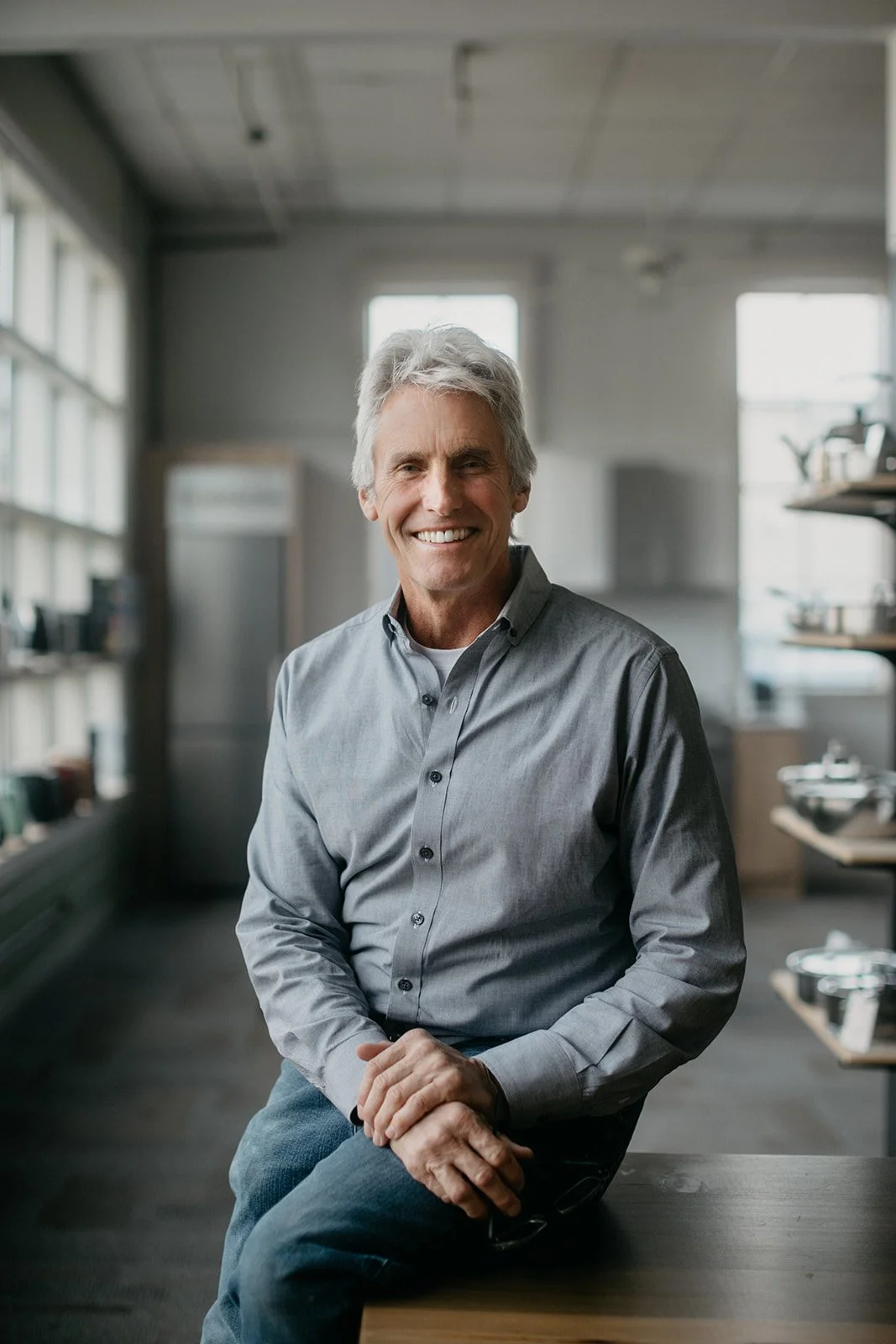 A smiling older man with gray hair, wearing a gray button-down shirt, sitting on a wooden table in a well-lit kitchen or café with shelves in the background.