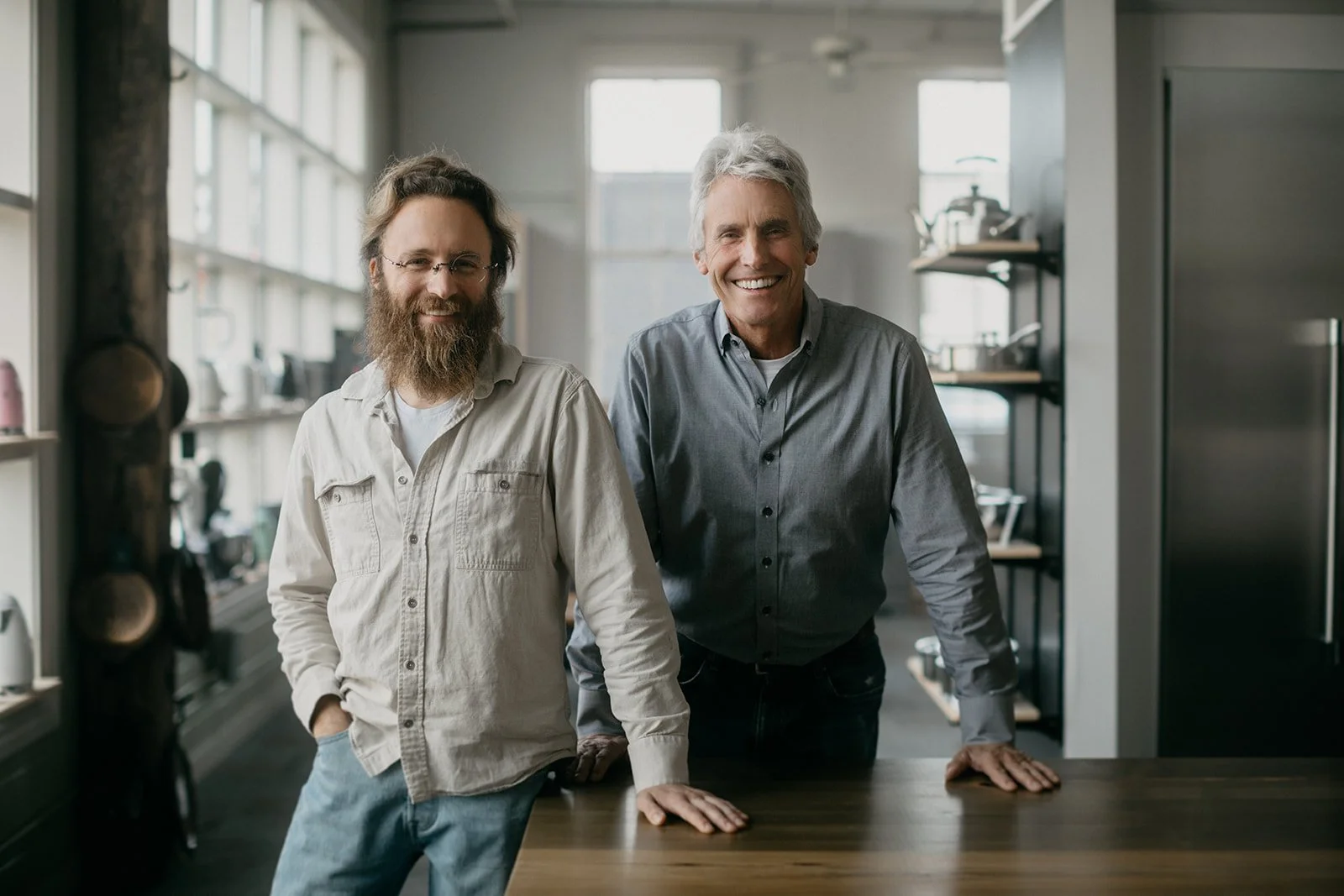 Two smiling men standing in a modern kitchen, leaning on a wooden table, with large windows in the background.