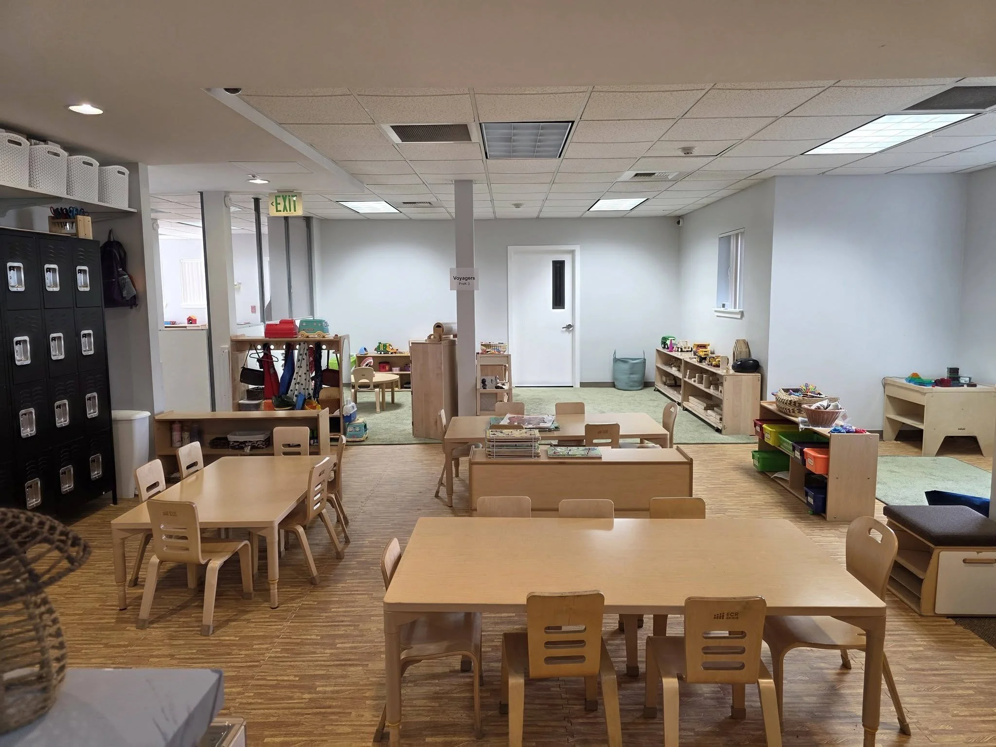 A classroom with small tables and chairs, storage lockers, activity shelves, and toys, indicating it is a preschool or kindergarten classroom.