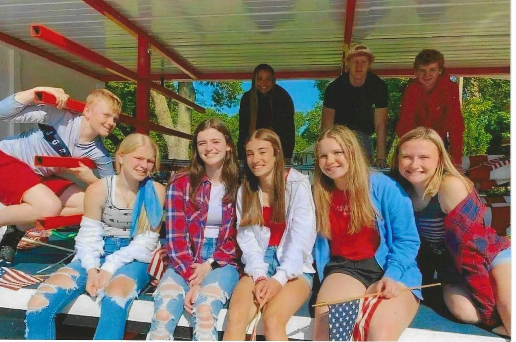Group of smiling teenagers sitting on a parade float, holding American flags, wearing casual summer clothes.