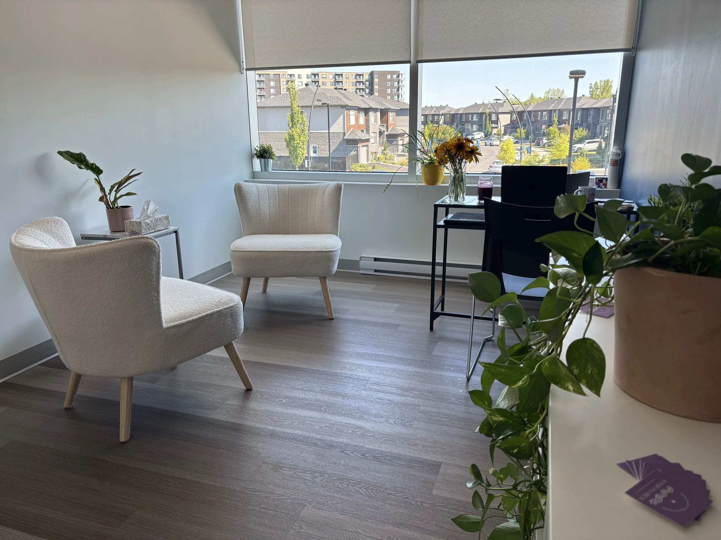 Coin salon avec deux fauteuils blancs, une table avec une plante verte, une grande fenêtre avec vue sur des bâtiments résidentiels et végétation, une plante en pot sur une surface