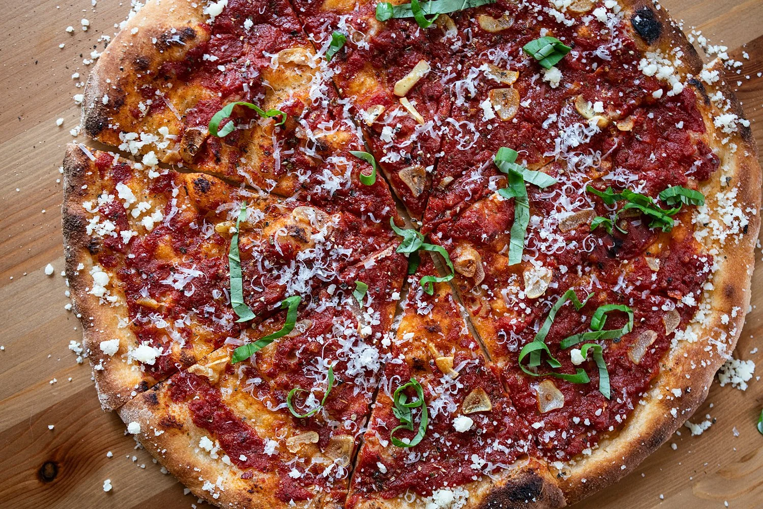 A close-up view of a cooked pizza topped with tomato sauce, shredded cheese, sliced garlic, chopped green onions, and herbs, sitting on a wooden surface.