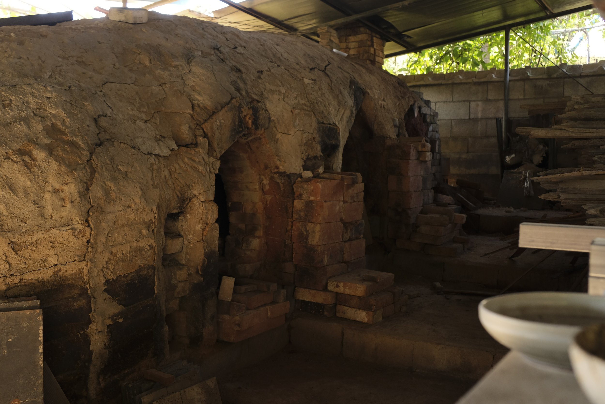 A rustic brick and mud oven in a workshop or storage area, with a ceramic bowl in the foreground.