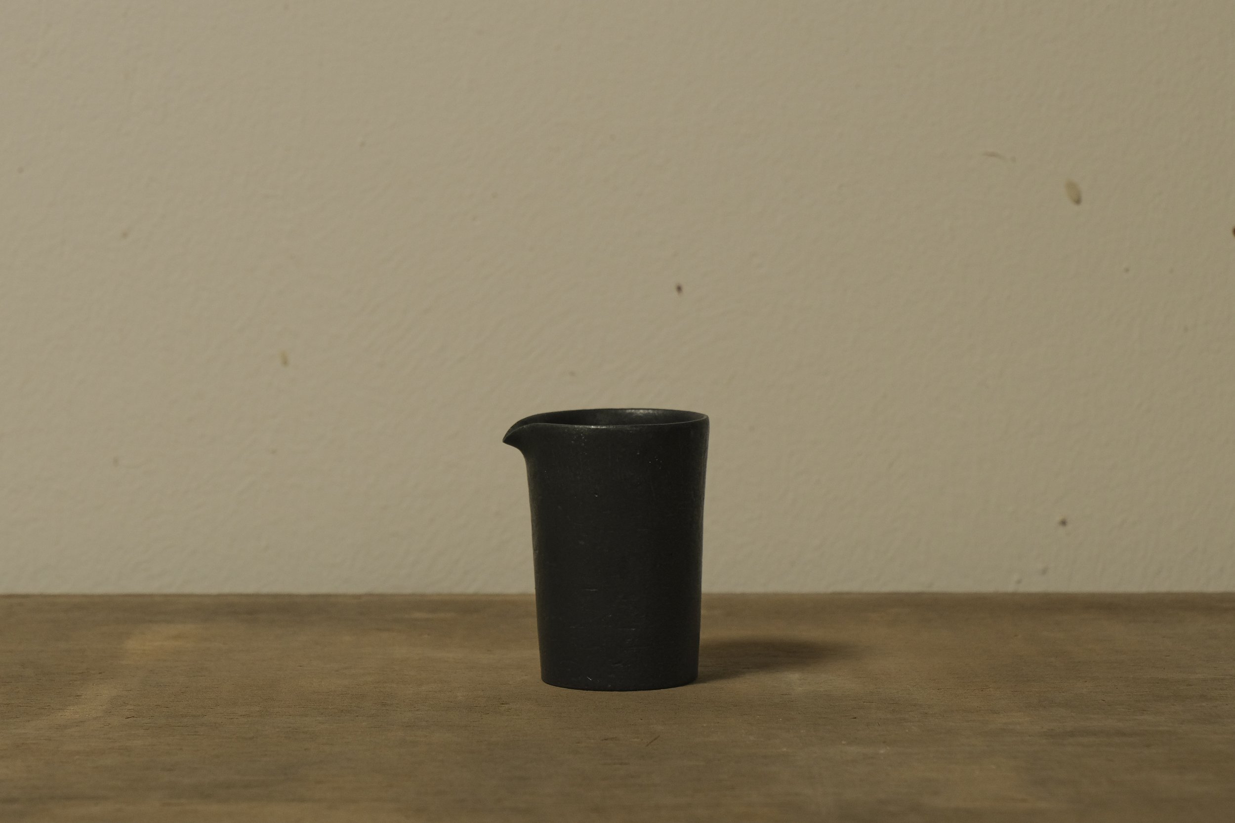 A small, black ceramic pitcher with a spout, placed on a wooden surface against a plain, beige wall.