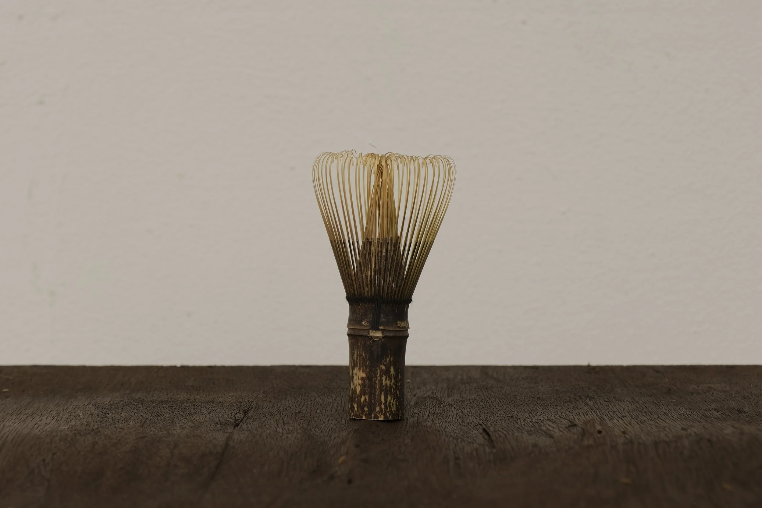 A traditional bamboo whisk for preparing matcha tea, placed on a wooden surface against a plain wall.