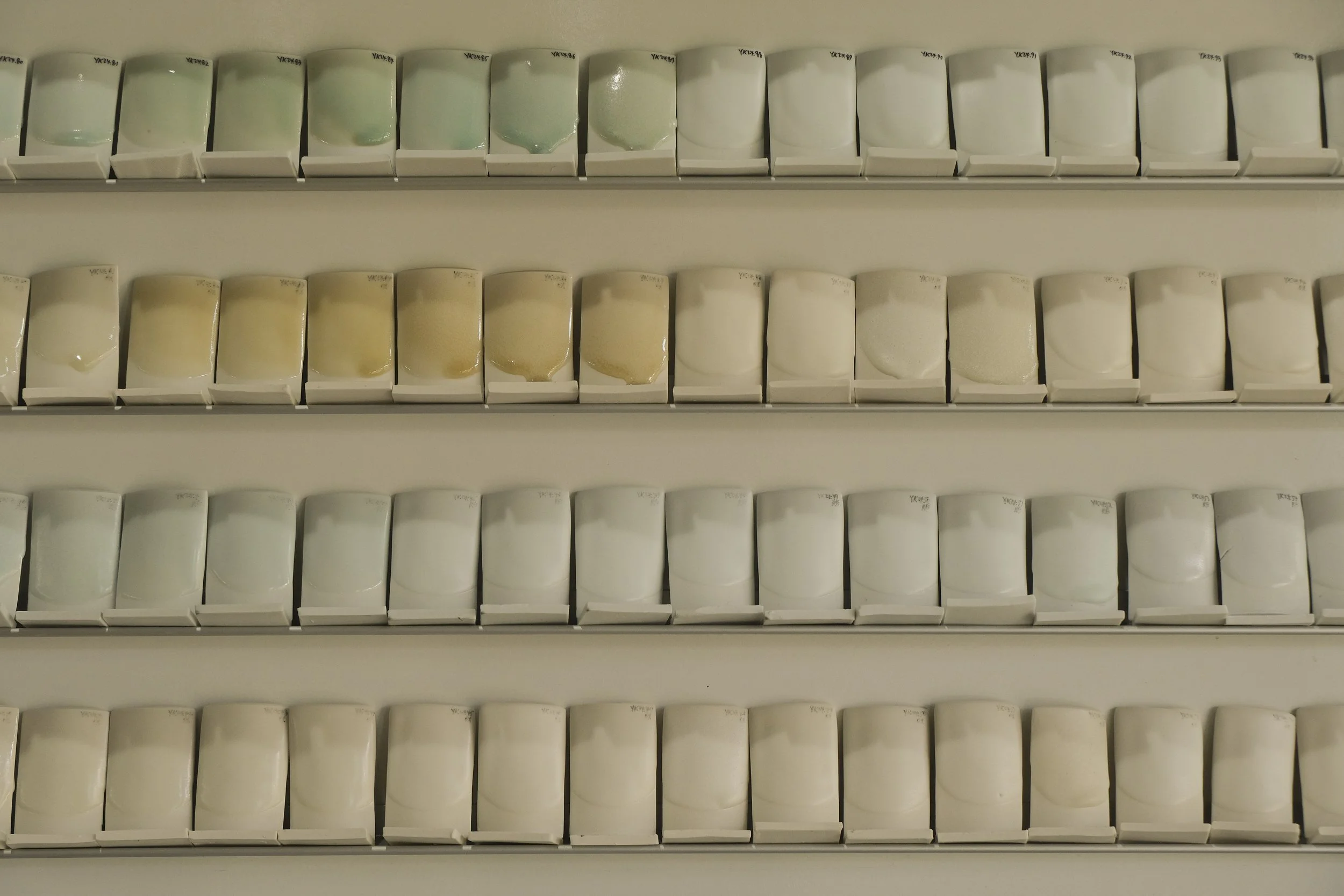 Display of multiple paint color samples on white shelves, arranged in three rows with varying shades of beige, white, and light pastel green.
