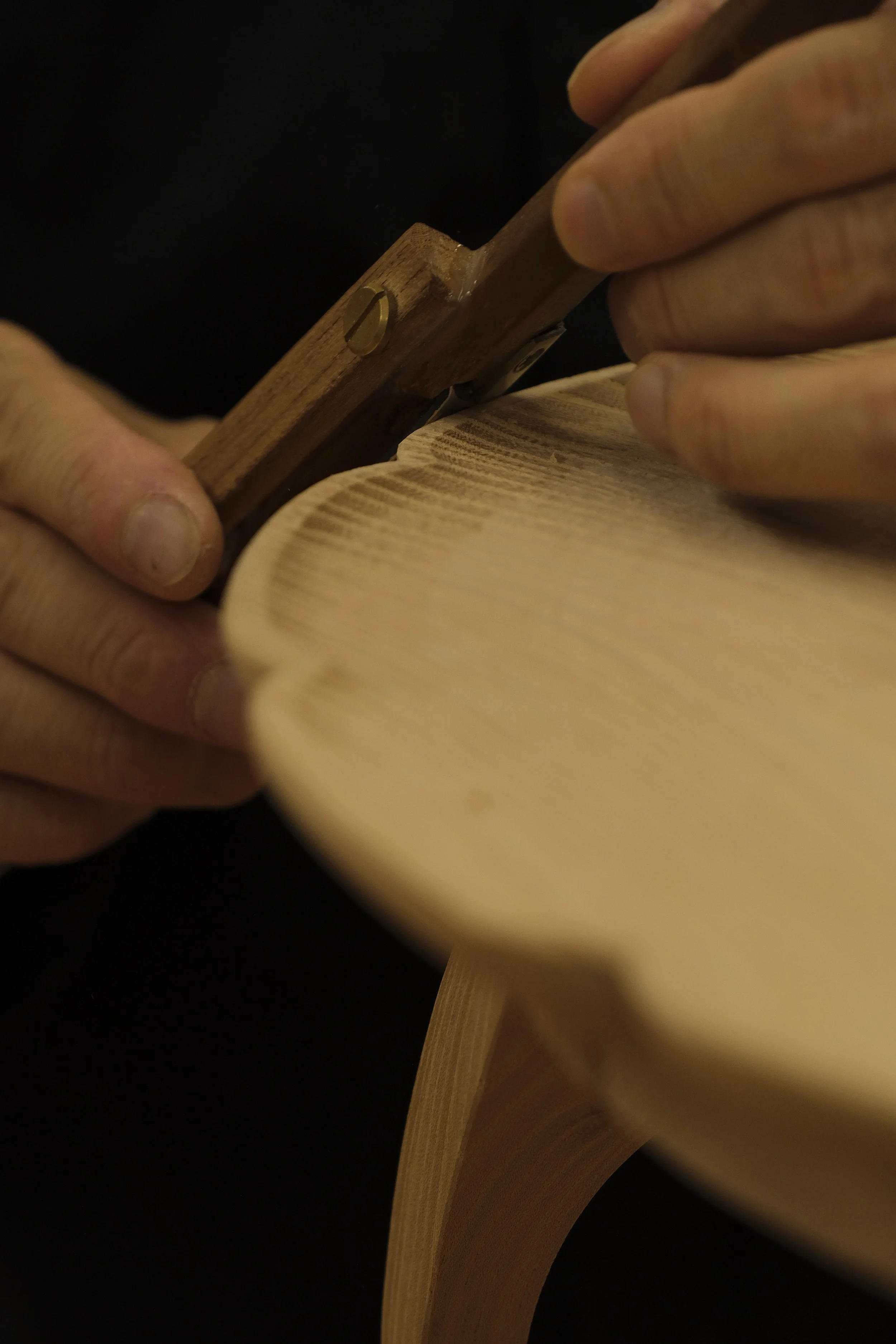 Close-up of a person using a hand plane to smooth the surface of a wooden piece, with a black background.