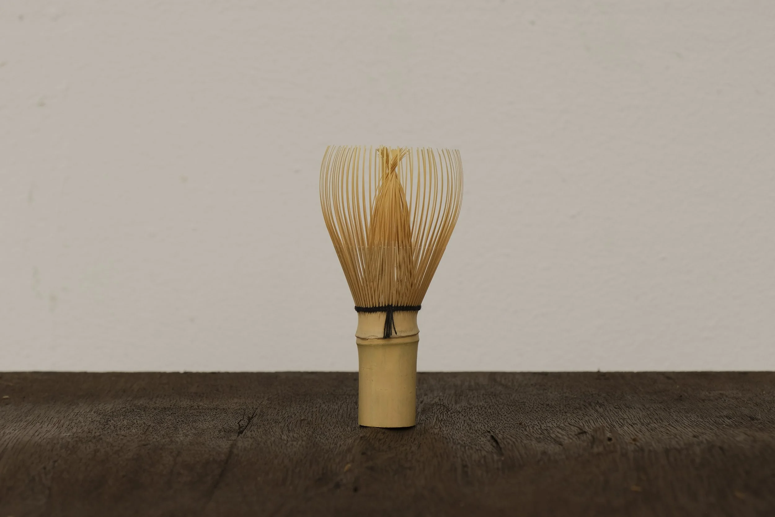A traditional bamboo tea whisk with thin loops of bamboo, standing upright on a wooden surface against a plain light-colored wall.