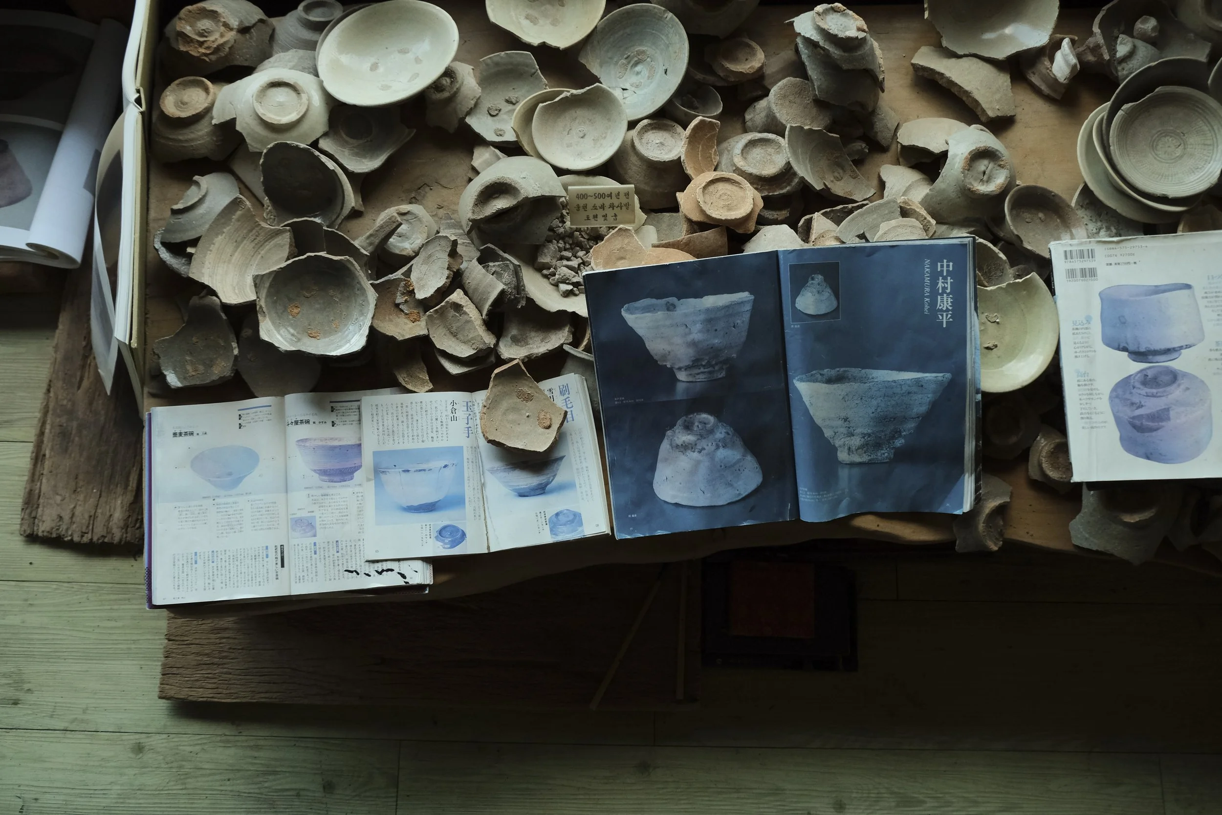 Collection of ancient pottery shards and broken ceramic bowls on a wooden table, with printed reference books and magazines about pottery.