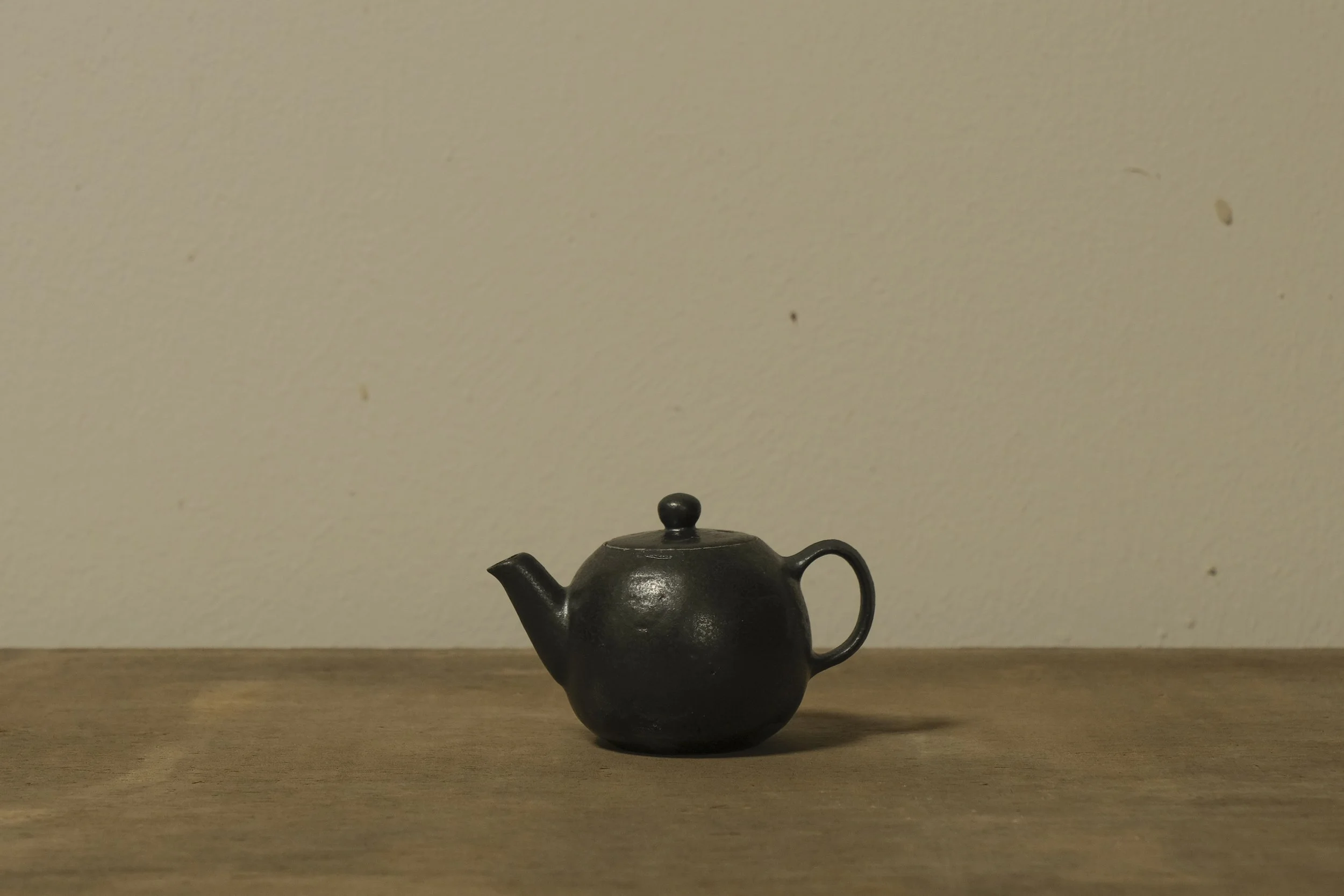 Small black teapot on wooden surface with plain beige wall background.