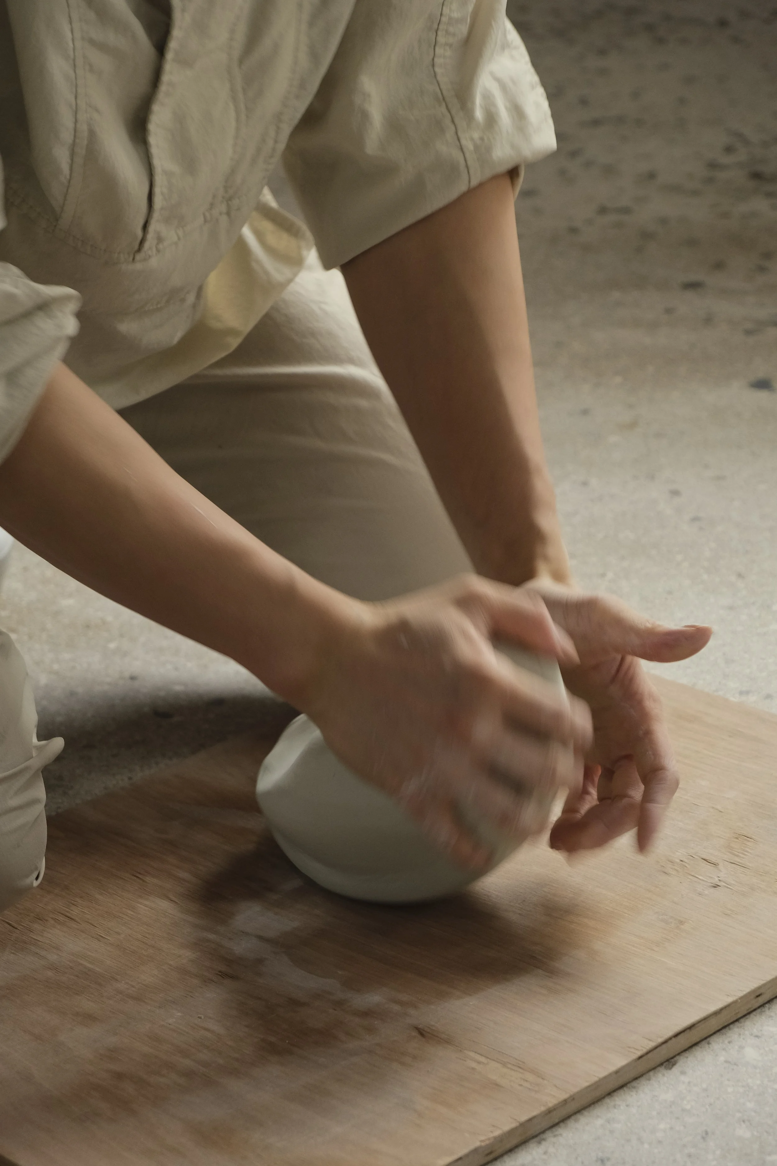 A person kneeling on the ground, shaping a clay vessel on a wooden board.