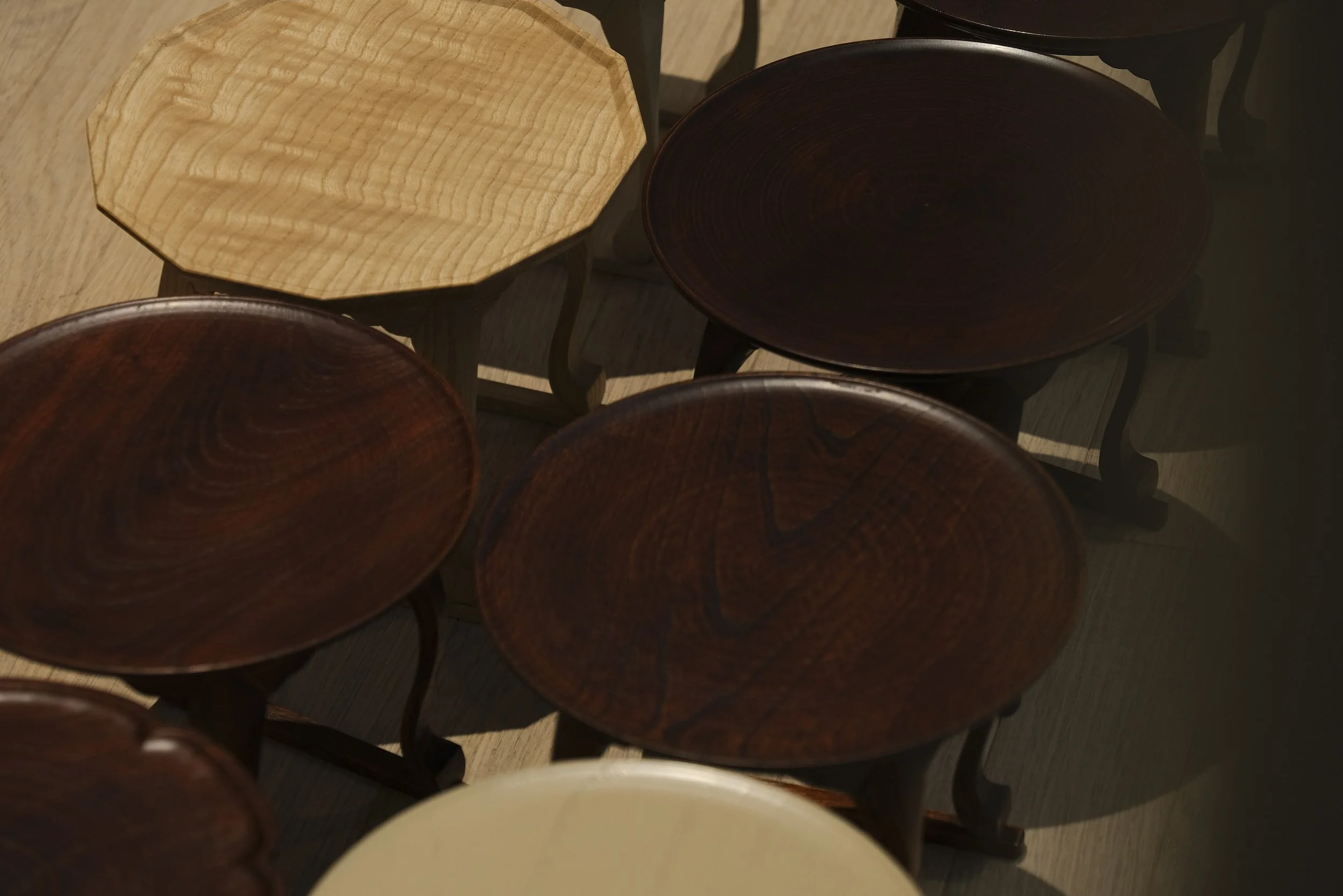 Various wooden stools with round seats, featuring light and dark wood tones, arranged closely in a room with wooden flooring.