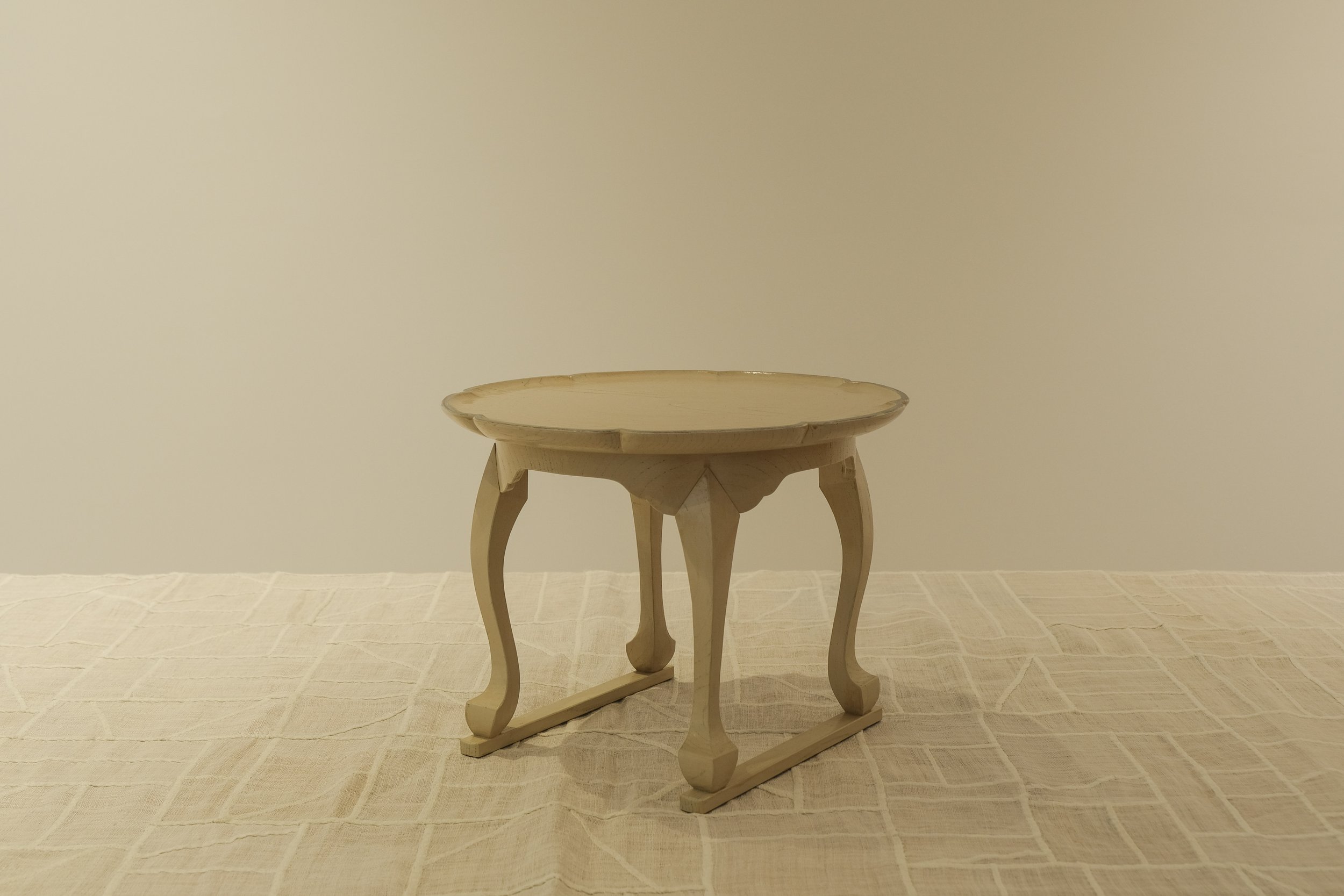A small, ornate wooden table with curved legs and a round top, placed on a beige checkered tablecloth against a plain, light-colored wall.
