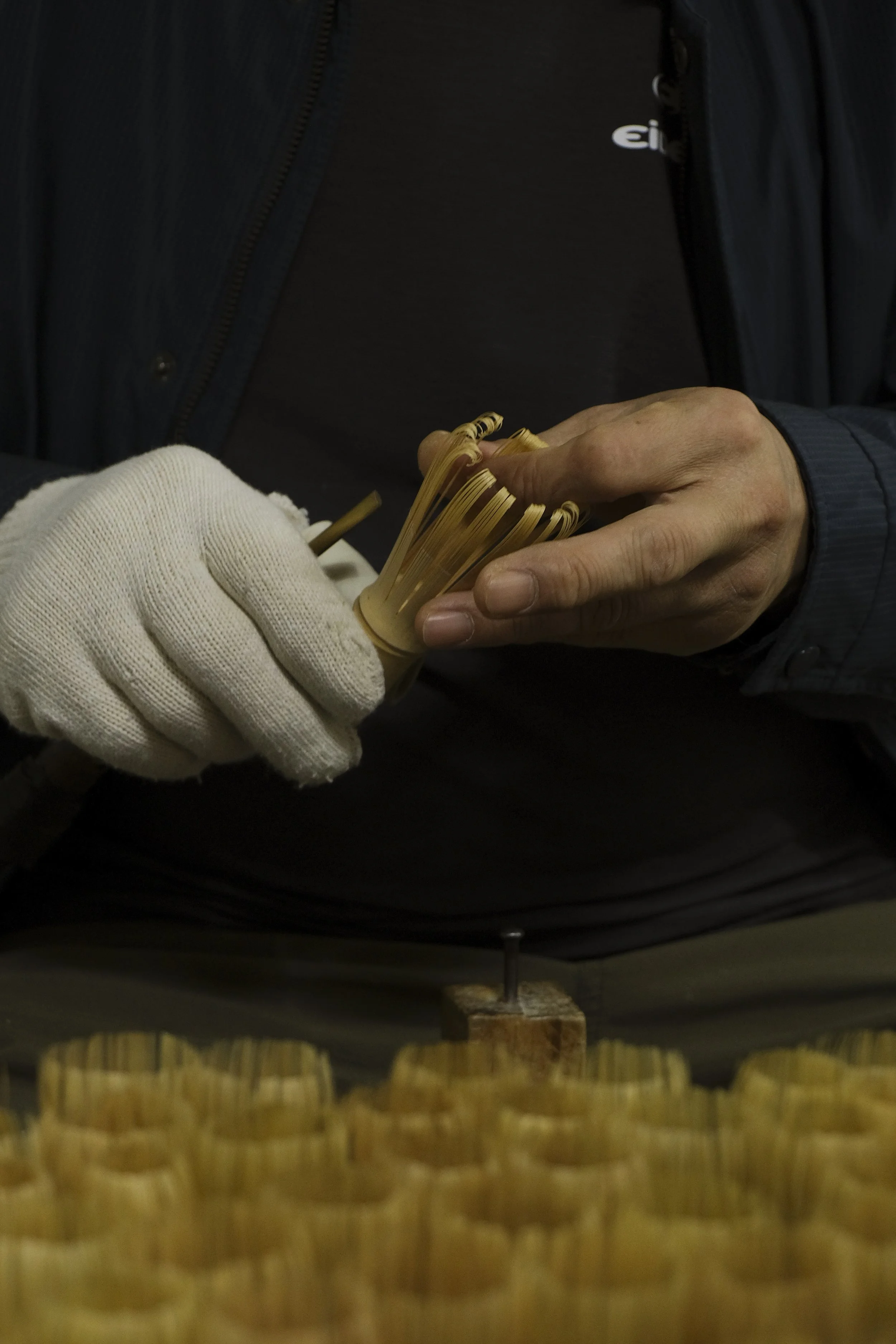 Close-up of a person assembling or shaping dried pasta by hand, wearing a glove on one hand and a dark jacket.