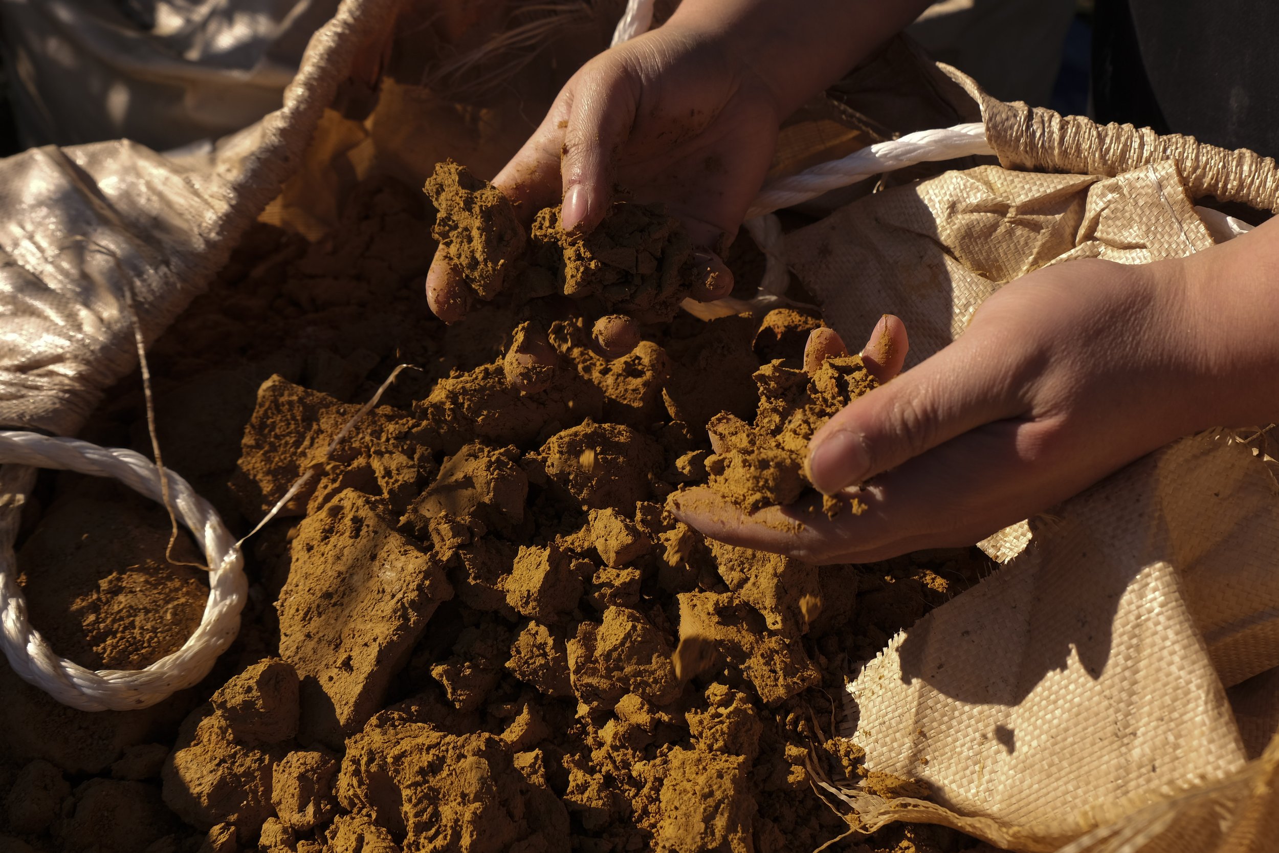 Hands holding and examining clumps of orange-brown soil or clay outdoors, with a woven sack and rope in the background.