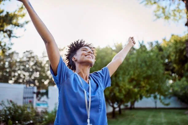 A woman in scrubs smiling with arms raised outdoors during daytime.