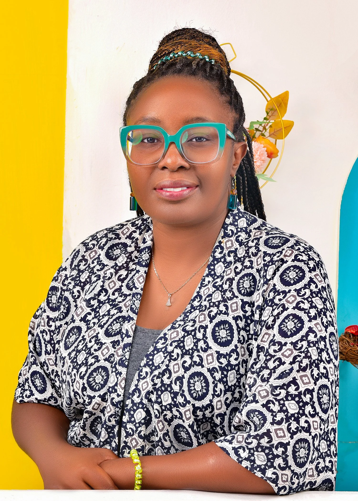 A woman with glasses, braided hair with beads, and colorful jewelry, wearing a patterned shirt, standing against a vibrant, colorful background with yellow, white, and blue sections and decorative floral art.