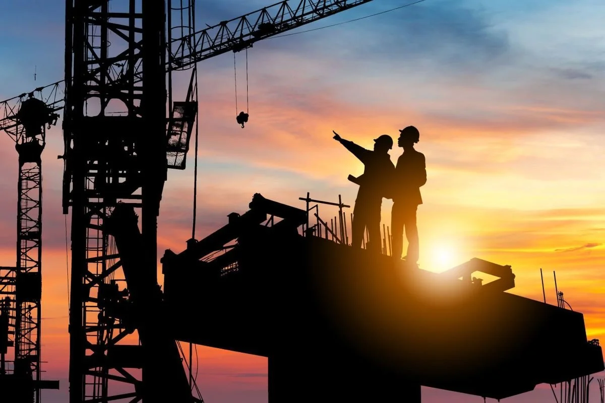 Silhouettes of two construction workers on a site during sunset, with a large crane nearby.
