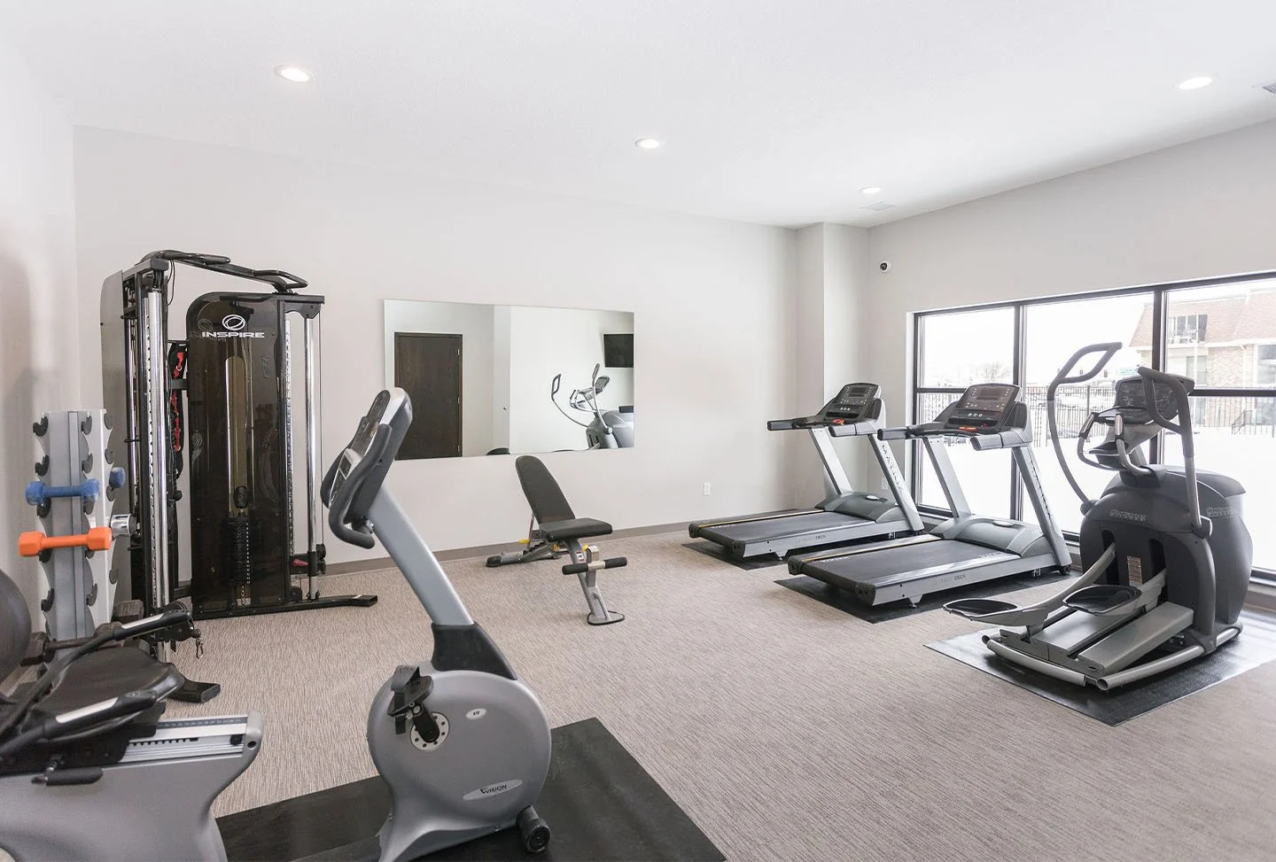Workout room with treadmill, elliptical, weight machine, stationary bike, and free weights, bright natural light.