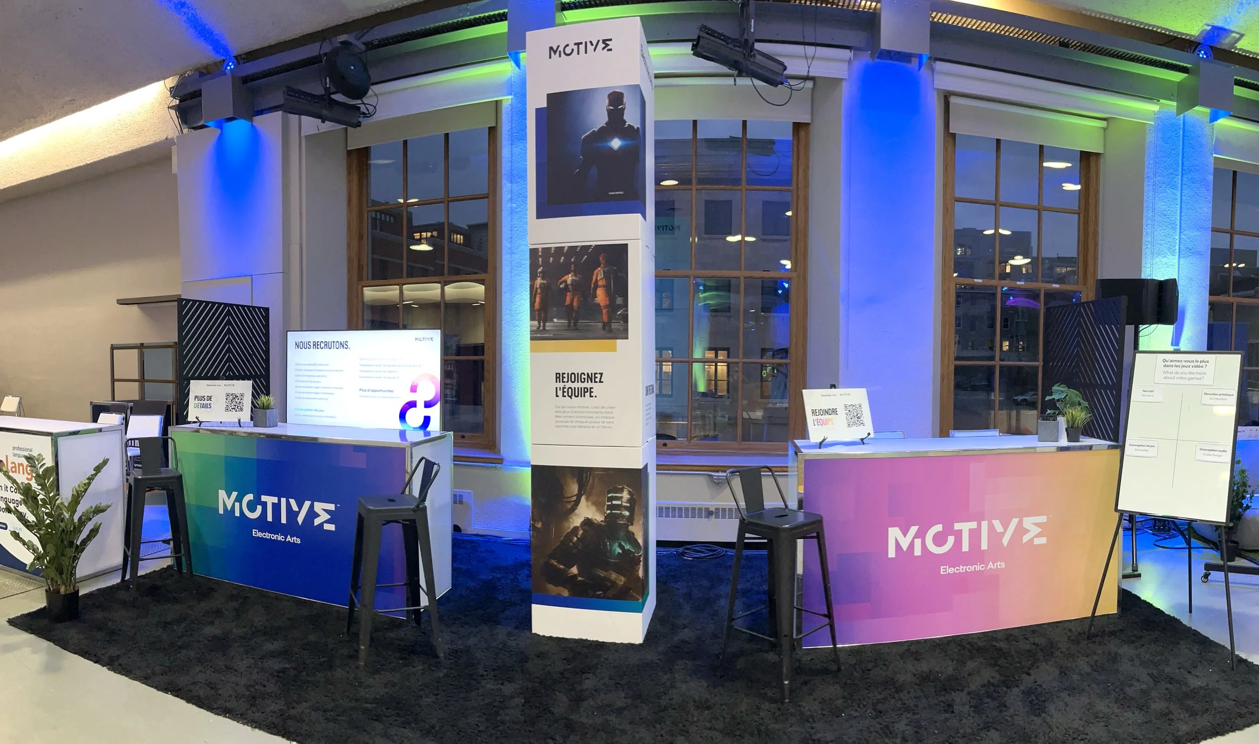Exhibition booth for 'MOTIVE' with colourful branding, two counters, chairs, large windows, promotional banners, digital screens, and informational signs about recruitment and team joining, illuminated with blue and green lighting.