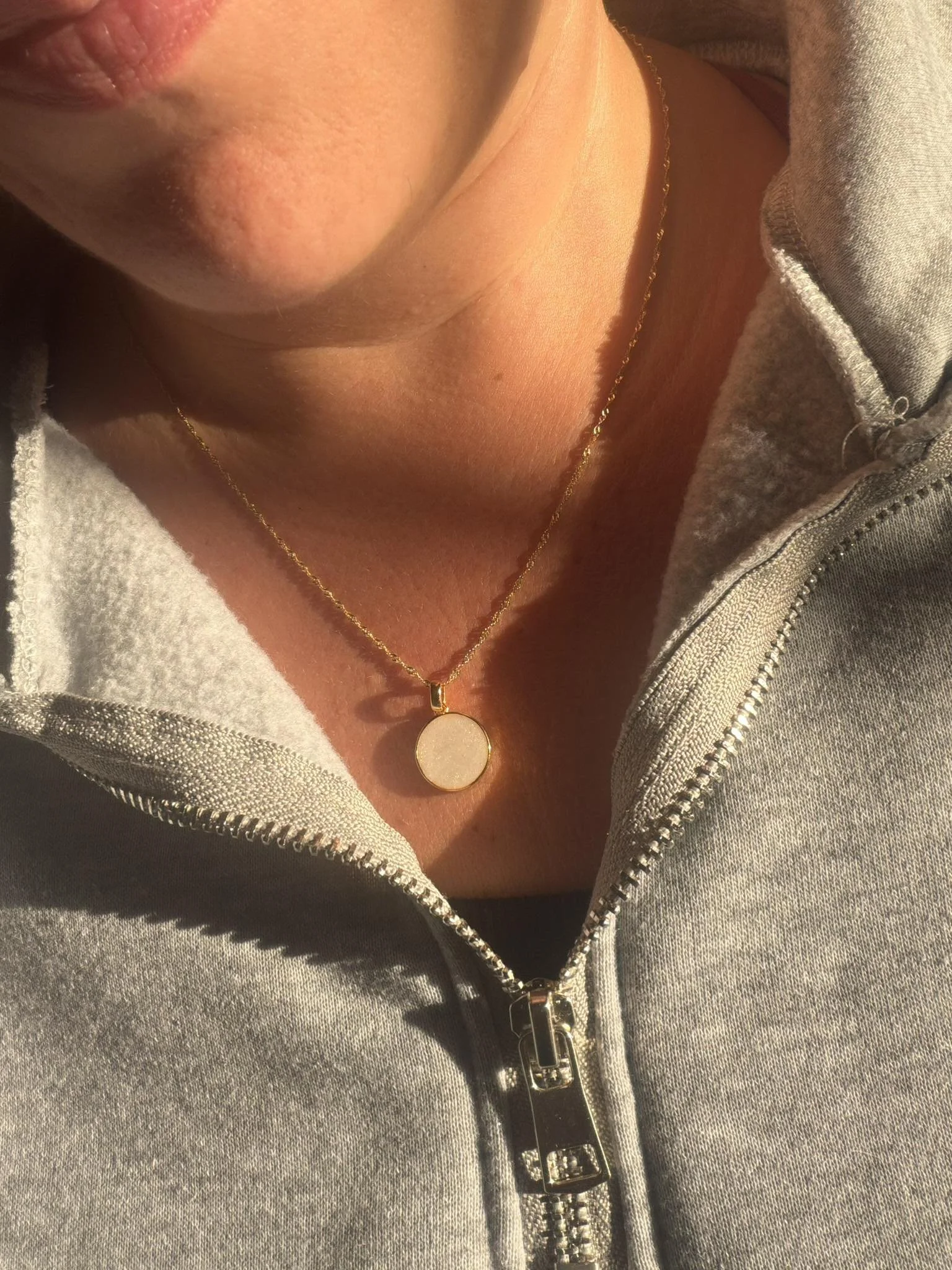 Close-up of a person's neck and chest showing a gold chain necklace with a round pendant, wearing a gray zip-up hoodie.