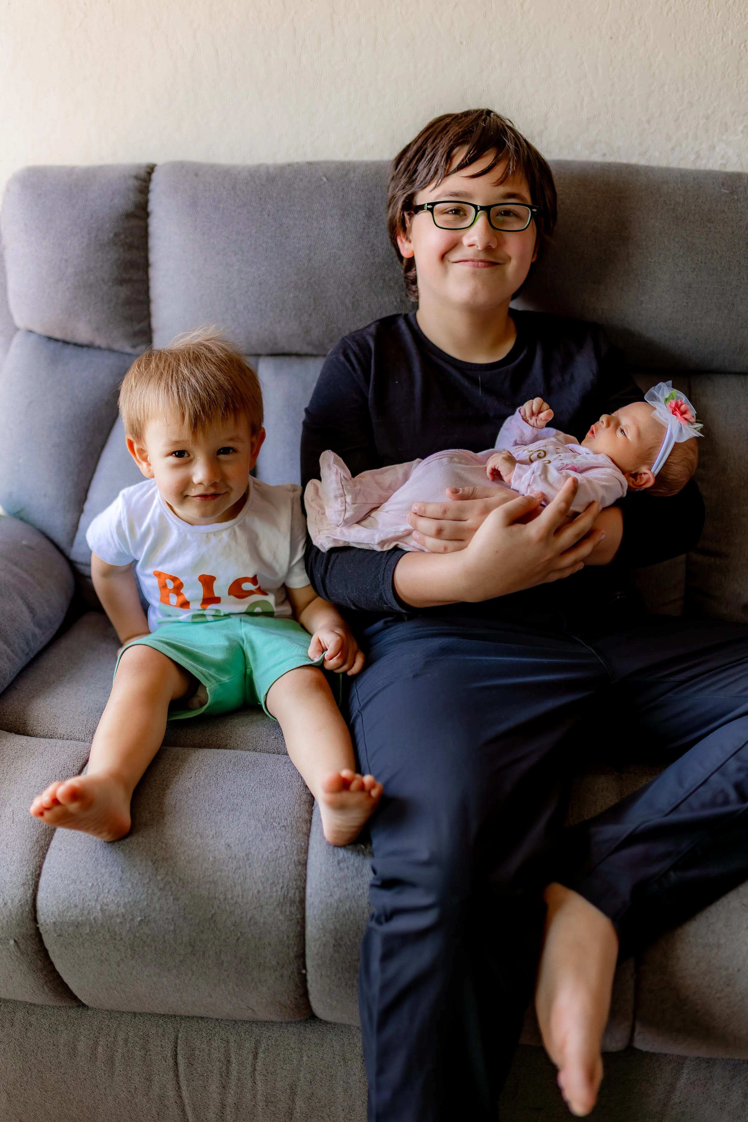 A boy with glasses holding a baby girl on his lap while sitting on a gray sofa, with a younger boy sitting next to him, all smiling.