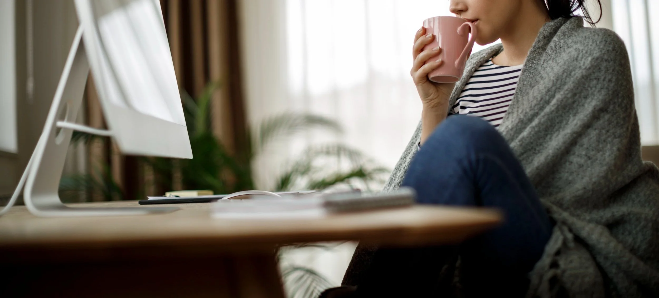 Woman sitting at a desk, drinking from a pink mug, with a computer monitor in front of her in a well-lit room.