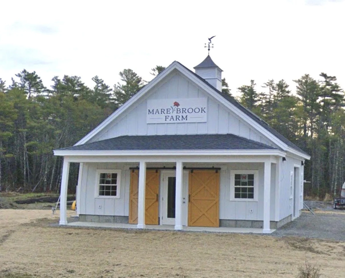 White barn-style building with a sign that reads 'Mareybrook Farm', wooden double doors, and two small windows, set in a rural area with trees in the background.