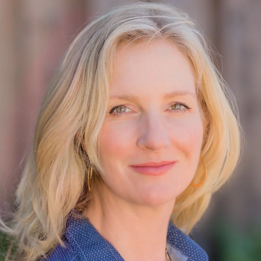 Close-up portrait of a woman with blonde hair, piercing blue eyes, and a light complexion, smiling softly outdoors with a blurred background.