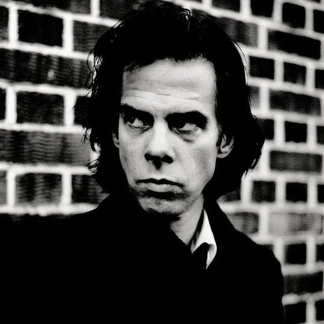 Black and white photo of a man with dark, wavy hair standing in front of a brick wall, looking to the side with a serious expression.
