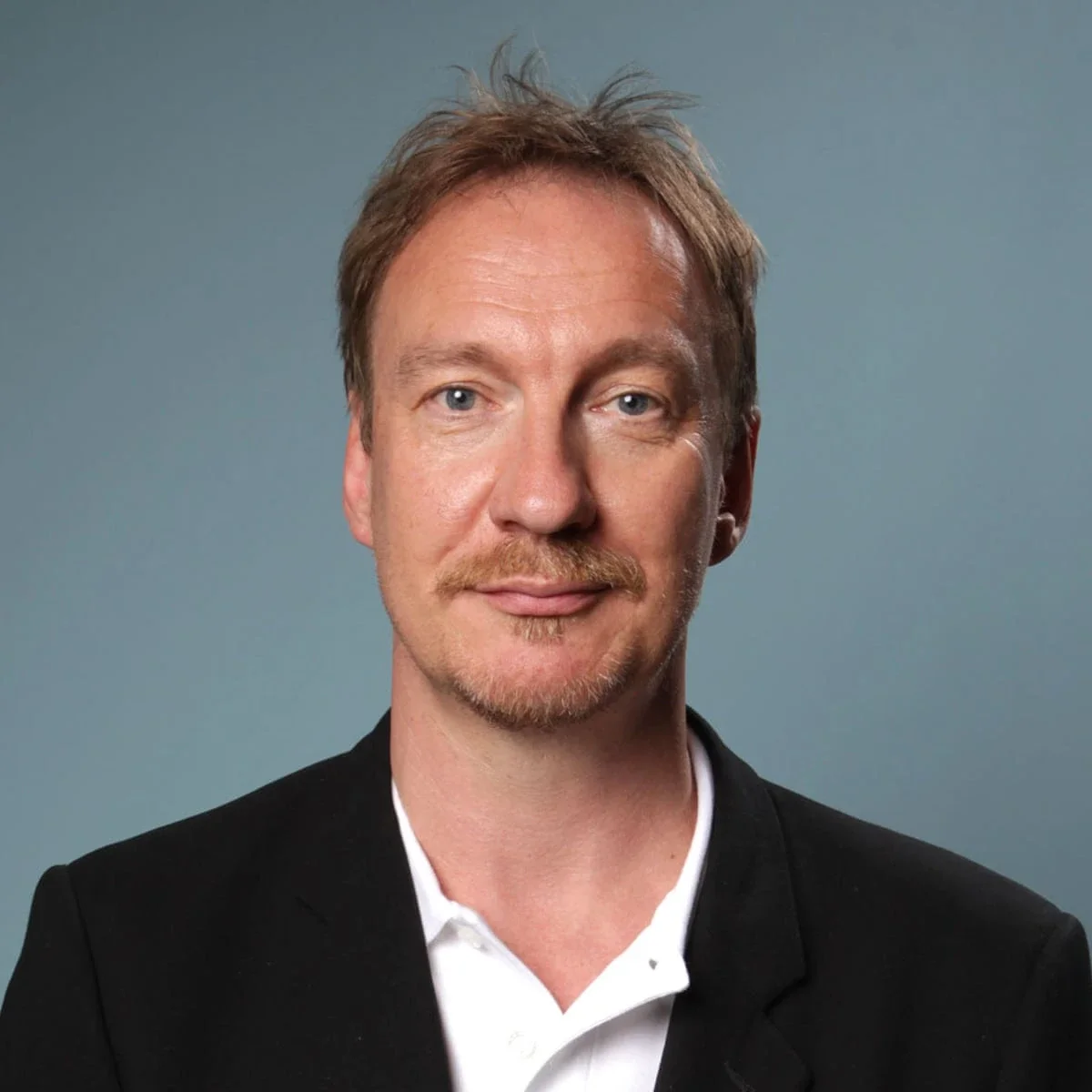 A professional headshot of a middle-aged man with light brown hair, blue eyes, a goatee, and a slight smile, dressed in a black blazer and white shirt against a plain grey background.