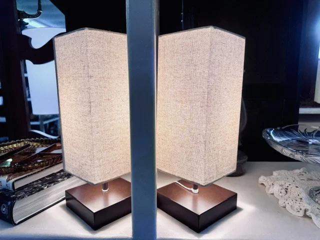 A pair of beige fabric lamps with dark base, turned on and placed on a table.
