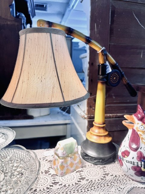Table lamp made from a vintage telephone, with a lampshade and a telephone receiver as part of the lamp's arm, surrounded by decorative items like a ceramic fish and a glass dish.