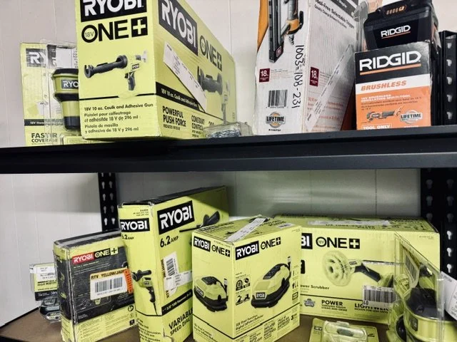 Shelf displaying various RYOBI power tools and accessories in yellow and black packaging, along with RIDGID tools to the side.