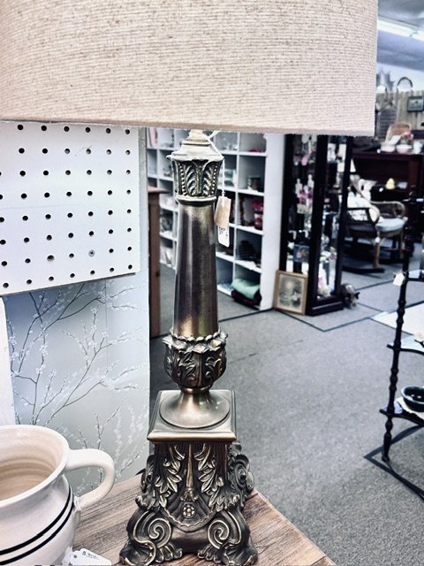 A decorative table lamp base with ornate detailing, placed on a wooden surface in an antique shop.