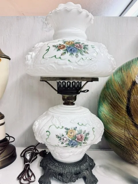 Vintage white porcelain table lamp with floral design, ornate base, and scalloped top, situated on a surface with other decorative items.