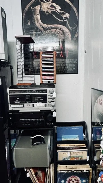 A vintage stereo system and record player in a room with a "Kiss the Dragon" movie poster on the wall behind them. There are vinyl records in a rack and a collection of cassette tapes.