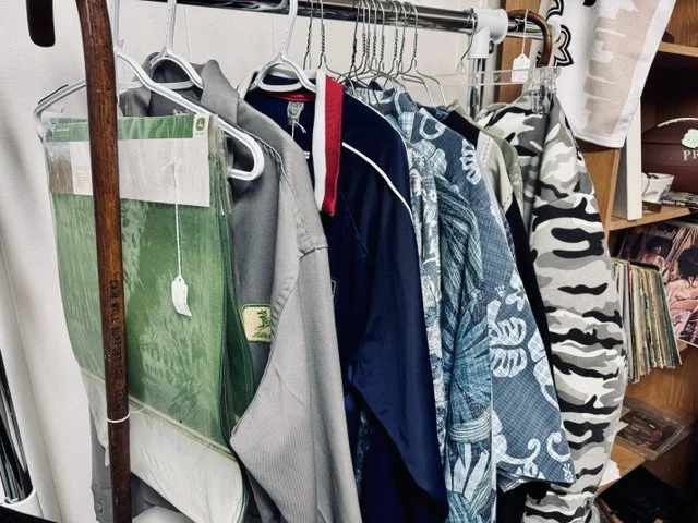 Clothes hanging on a metal rack, including jackets, hoodies, and shirts, with a cardboard box and books in the background.