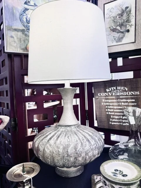 Table lamp with a large, textured, round ceramic base and a white lampshade, placed on a black table with decorative items and artwork in the background.