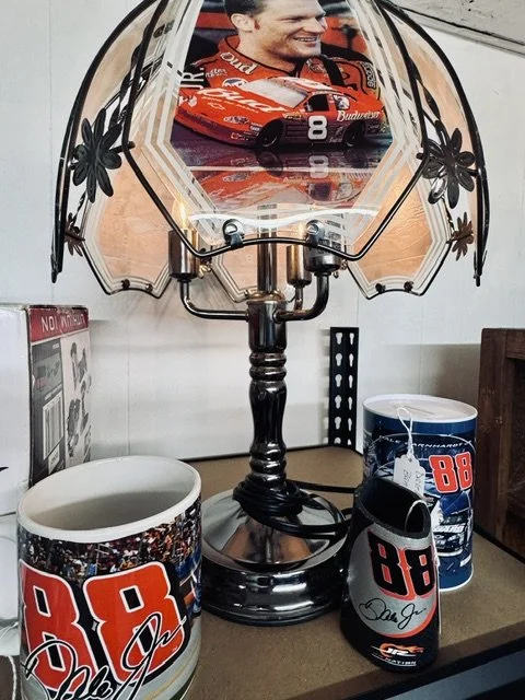 Table lamp with a shade featuring a racing car and a smiling racetrack driver, flanked by two coffee mugs and a travel mug, all decorated with the number 88, NASCAR logo, and racing colors.