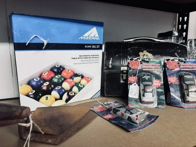 Box of billiard balls, a black grill, and two packages of toy cars on a shelf.